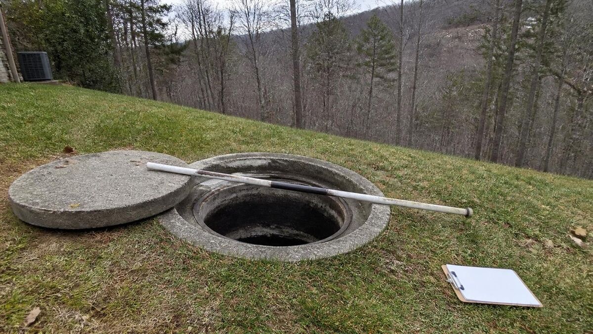 An open concrete septic tank access hole on a hillside lawn, the lid set aside and a measuring pole across the opening